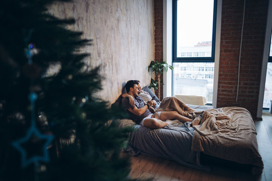 Two attractive guys watching their romantic christmas photos on smartphone, resting in bed, embracing each other. - Powered by Adobe