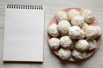 Homemade white mexican wedding cookies on pink plate, blank notepad over white wooden surface, overhead view. Copy space.
