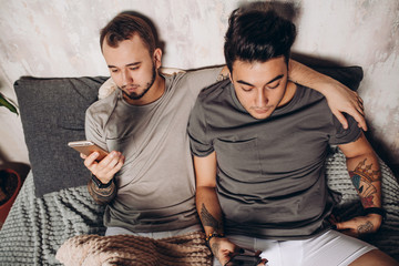 Alternative relationships, couple of young gay guys lying together in bed and using smartphone to...