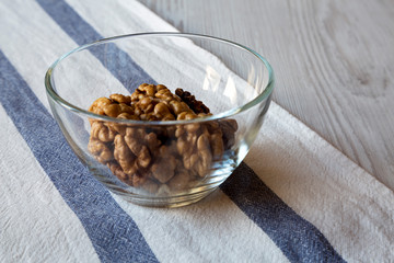 Walnuts in glass bowl on cloth over white wooden background, closeup.