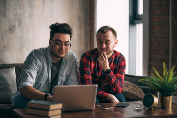 Romantic partners in love and business. Caucasian amused gay couple looking at laptop screen while resting in living room with loft interior. Dominant man wearing spectacles and and using laptop