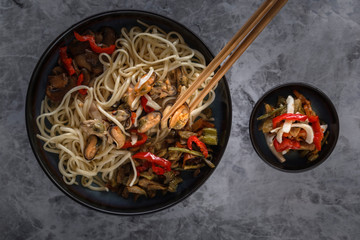 Traditional wooden chopsticks with coiled noodles on the background of a dish with seafood, vegetables and peppers. Close-up. Top view