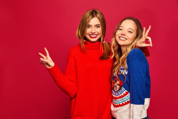 Two beautiful smiling gorgeous girls looking at camera. Women standing in stylish winter warm sweaters on red background. Christmas, x-mas, concept.