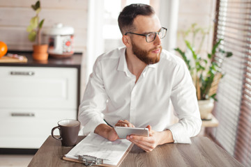 Young confident male examines house purchase agreement papers, buying the house, using smartphone...
