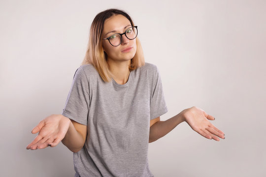 I don't know concept. Young laughs woman who do not understand what is happening. The girl in big eyeglasses and gray shirt raises hands and shrugging shoulders on a gray background copy spac
