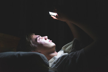 Depressed teenager browsing the internet on his mobile phone as he is lying on his bed in the dark. - obrazy, fototapety, plakaty