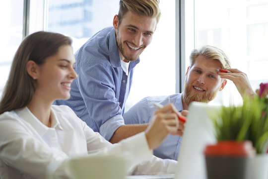 Group of young modern people in smart casual wear pointing at laptop and smiling while sitting in the office.