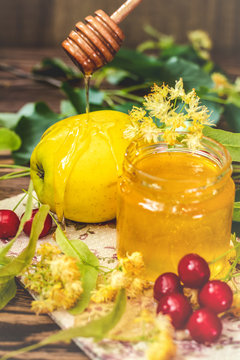 Open glass jar of liquid honey, apple and honey dipper, bunch of linden flowers
