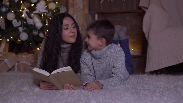 Mom with long hair reads a book to her son and kisses him lying on the floor near the Christmas tree on Christmas day.HD