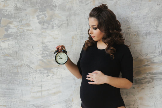 Portrait of beautiful pregnant woman in black bodysuit. She hold the clock in hands symbolic hinting at the remanining time befour the baby in born. Woman look at the clock