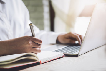 Study through technology, people use computer laptop and notebook with pen to take notes from work on wood desk.
