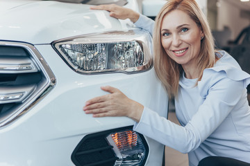 Mature businesswoman looking excited, hugging her new automobile, smiling joyfully to the camera. Female driver choosing new car at the dealership. Happy woman shopping for an auto. Ownership concept