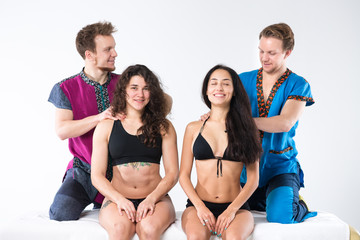 Theme massage and body care. Two male Caucasian twin brothers in uniform doing shoulder massage to two beautiful sexy European women in lingerie sitting on massage table ga isolated white background