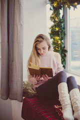 young blond woman in leggings sitting on the window. to read a book. christmas interior