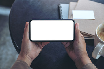 Top view mockup image of hands holding and using a black mobile phone with blank screen horizontally for watching with coffee cup and notebooks on table