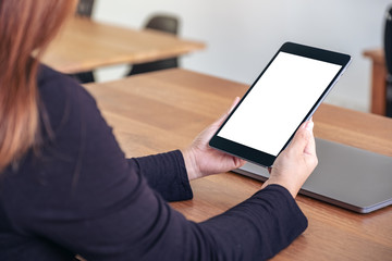 Mockup image of hands holding and using black tablet pc with blank white desktop screen with notebook on wooden table in office