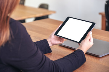 Mockup image of hands holding and using black tablet pc with blank white desktop screen with notebook on wooden table in office