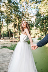 Fototapeta na wymiar Follow me my love concept. Attractive young woman dressed in white wedding dress holding hand of her boyfriend and walking on the beautiful garden to happy future.