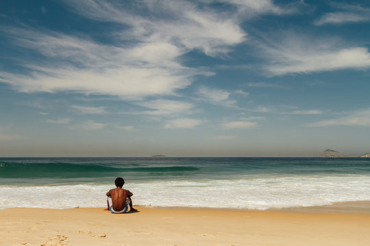 Young African American man sitting alone facing the sea on Copacabana beach in Rio de Janeiro, Brazil