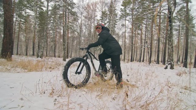 Professional extreme sportsman biker stand a fat bike in outdoors. Cyclist recline in the winter snow forest. Man walk with mountain bicycle with big tire in helmet and glasses. Slow motion in 60fps.