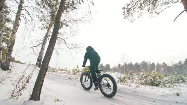 Professional extreme sportsman biker riding fat bike in outdoors. Cyclist ride in winter in snow field, forest. Man does trick on mountain bicycle with big tire in helmet, glasses. Slow motion 60fps.
