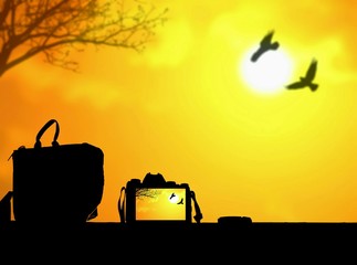 Silhouette digital camera and bag on tabletop is recording with blur background of birds are flying in sunrise sky in travel concept, illustration mode