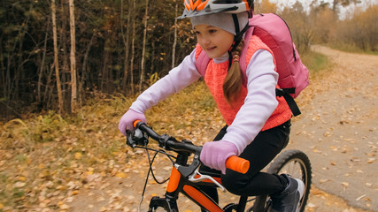 Fototapeta na wymiar One caucasian children rides bike road in autumn park. Little girl riding black orange cycle in forest. Kid goes do bicycle sports. Biker motion ride with backpack and helmet. Mountain bike hardtail.