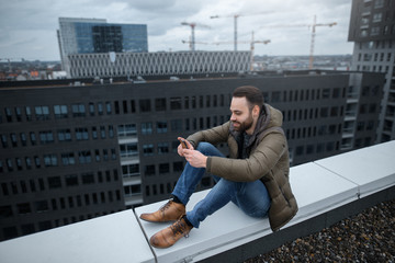 Smiling and bearded hipster guy looks at the social network in the smartphone, on the roof of a tall building.