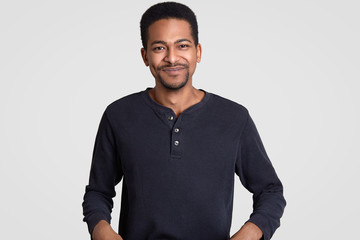 Photo of satisfied dark skinned guy with stubble, feels happy and pleased, being in good mood, wears casual clothes, models against white background. Afro American hipster smiles gently, stands indoor