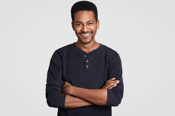 Indoor shot of handsome satisfied black man with arms folded, wears casual clothes, smiles gently, has Afro haircut, has pleasant talk with interlocutor, isolated over white wall. Positive emotions