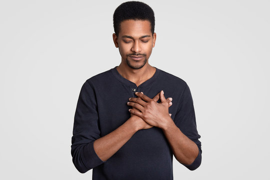 Photo of black man with faithful expression, keeps hands on chest, expresses sympathy, closes eyes, feels gratitude, dressed in sweater, models against white background. Body language concept