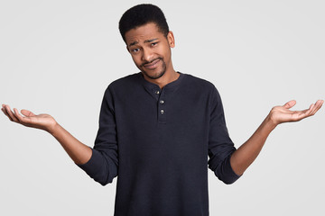 Cluless hesitant guy with dark skin, shruggs shoulders in apathy, feels doubt, has indecisive facial expression, wears casual outfit, isolated over white studio wall. People and hesitation concept