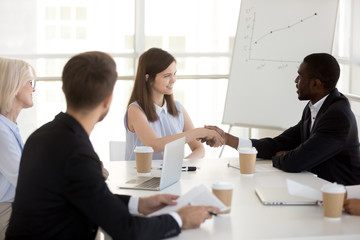 Smiling millennial caucasian businesswoman and african partner shake hands at office group meeting, friendly diverse employees handshaking greeting, welcoming, business acquaintance, respect concept
