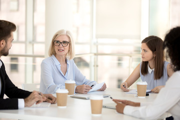 Serious mature team leader executive speaking at office briefing discussing paperwork report with young employees group, negotiating, giving instruction, delegating work, coaching and mentoring