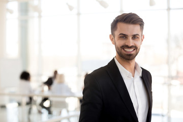 Obraz premium Smiling millennial businessman in suit looking at camera posing in office, portrait of happy confident male ceo, successful entrepreneur, executive leader, company manager, business coach headshot