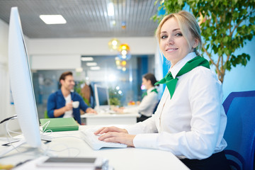 Smiling beautiful young lady wearing white blouse and neck scarf sitting at table and tying on computer keyboard while working in bank and filling online customers card. - obrazy, fototapety, plakaty
