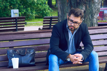 Young man with sad facial expression sitting on a bench in the park. Office worker lost his job....