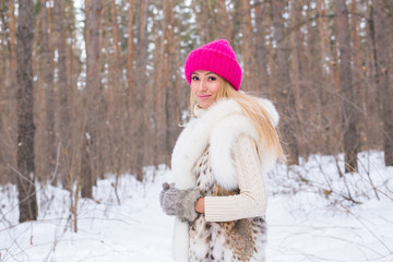 Fashion, season and people concept - Happy blonde woman in a pink hat and gloves and sweater in the winter forest is smiling