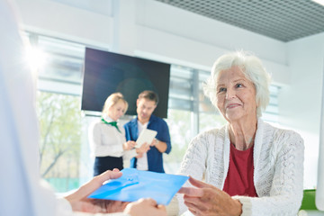 Positive attractive senior lady customer sitting at table and looking at personal manager while receiving envelope with documents in bank