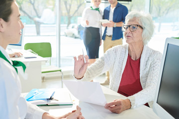 Displeased strict senior lady client in glasses sitting at table and making stop gesture to manager...