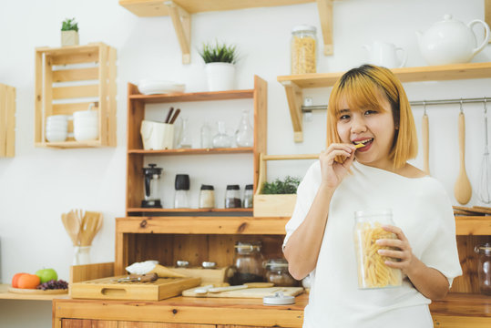 Asian woman holding and eating junk food snack and chips in her kitchen at home, female enjoy unhealthy food. Lifestyle women relax and enjoy junk food in kitchen at home concept.