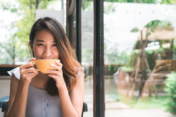 Cheerful asian young woman drinking warm coffee or tea enjoying it while sitting in cafe....