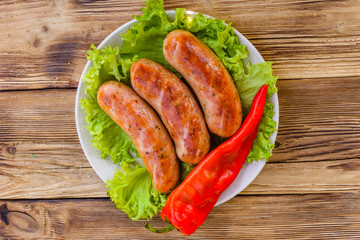 Ceramic plate with grilled sausages, pepper and lettuce leaves on wooden table. Top view