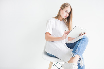 Portrait of a young stylish blonde girl in a white T-shirt and blue jeans using a tablet on a white background
