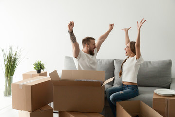 Happy excited young couple sitting together on couch, sofa celebrating moving at new house with cardboard boxes with belongings, family just arrived in new home, man and woman start living together