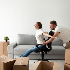 Carefree excited young couple having fun together playing with office chair in living room while unpacking boxes, man pushing chair woman riding on, family buy new house looking forward to good future