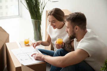 Young smiling couple discussing, pointing at house architectural plan, drinking juice, sitting together, planning new apartment interior design, remodeling, renovation, cardboard boxes with belongings