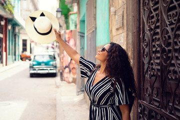 Young beautiful black woman with a typical cuban hat standing in the old streets of Havana Cuba in front of a classic car holding the hat up high.