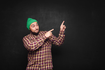 Young european man with beard in green knitted hat thinks, looks surprised and puzzled. Showing fingers upwards and right side. Black background, blank copy space for text or advertisement