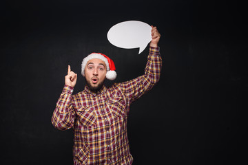 Funny serious  man in red Christmas Santa hat holds empty white cardboard as blank or mock up with copy space for text. Black background